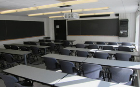 Photograph of an average-sized classroom with two large chalkboards covering the length of two sides of the room. Desks are long tables with movable chairs.