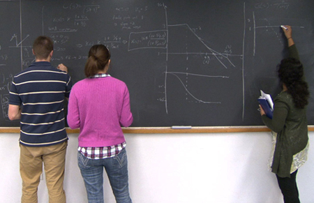Three students work at a long chalkboard to solve a recitation problem.