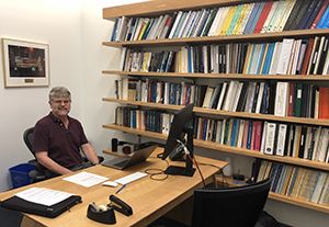 Man sitting at desk. Bookshelves with books to his left.