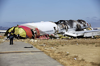 A crash investigator wearing an NTSB shirt approaches the burned-out body of a crashed passenger jet off the end of a runway.