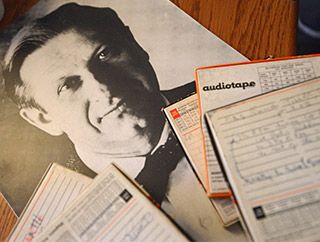 On a wooden tabletop, a black and white photograph of a distinguished-looking man wearing a bow tie rests beneath a pile of reel to reel tape boxes.