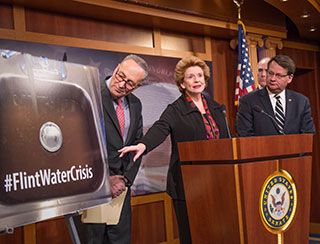 A red-haired, casually dressed woman, flanked by two suit-wearing gentlemen, stands at a podium and points to a large photo of a sink filled with dirty water and words reading: #FlintWaterCrisis.