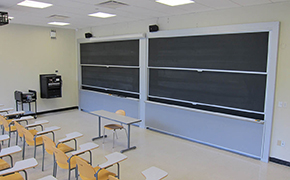 A well-lit, medium-sized room with a couple of rows of desks and chairs facing a small table positioned in front of two blackboards.