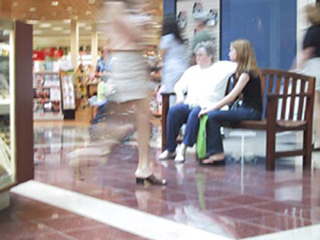 A photo of people sitting at a mall bench with people walking by.