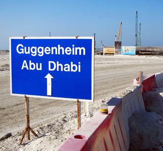 A photograph that features a large blue sign in the foreground that reads "Guggenheim Abu Dhabi." A construction site with cranes can be seen in the background.