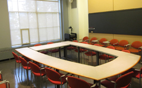 Classroom with 14 red chairs arranged around tables in a rectangular position. A blackboard runs along wall. Floor to ceiling windows.