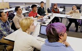 Seven students and an instructor sit at tables arranged in a u-shape. Two people have open laptops in front of them.