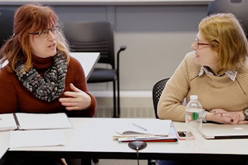 Two women sit side by side a classroom table. One of the women is speaking and gesturing, while the other looks in her direction.