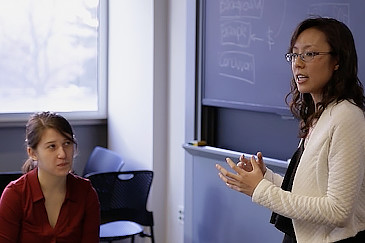 Woman with dark hair and black glasses stands in front of a blackboard; she speaks and gesticulates. A female teaching assistant is in positioned in the lower lefthand corner of the image; she looks toward the woman speaking.