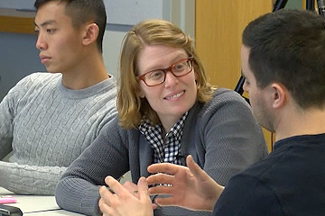 Woman with light hair and red glasses sits at a table between two male students. The woman looks toward the male student on her left. The male student gesticulates.