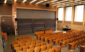 A photograph of a large tiered lecture hall consisting of rows of wooden seats and six sliding chalkboard panels at the front.