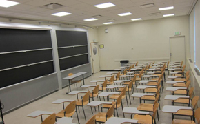 Classroom with 23 moveable desks with arm tablets facing two blackboards. A small table and chair are positioned in front of the blackboard. An overhead projector is to the far left of the room.