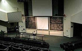 Lecture hall with green tablet armchairs arranged in tiered groups (capacity for over 100 students). Armchairs are facing the sliding chalkboards. Two tables are in front of the chalkboards. Three white screens are near the chalkboards.