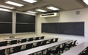 Classroom with narrow tables arranged in rows. Black chairs arranged behind each row of tables. Chalkboards on the front and side walls.