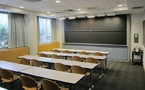 A classroom with a blackboard in the front, windows on the left, long tables pushed together to form desks and moveable chairs.