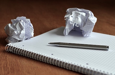 A photo of a notebook with a pen on top along with two balls of crumpled paper.