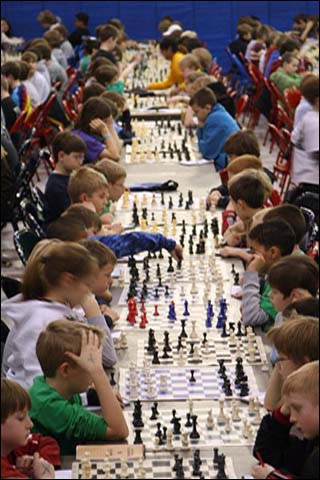 Dozens of children sit lined up at tables playing games of chess.