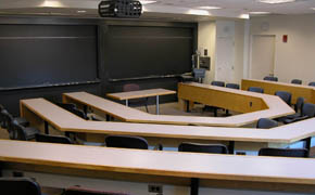 A photograph taken from the back of a classroom. The room has three rows of seating and blackboards at the front of the room.