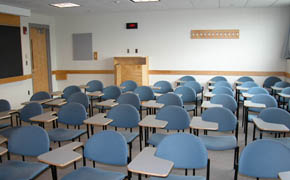 A photograph taken from the front of a classroom. The room contains several rows of chairs with desk arms.