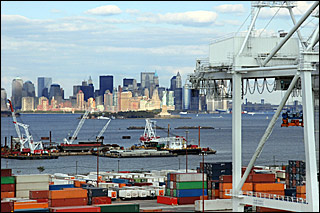 A photograph with the skyline of New York in the background and dozens of colorful shipping containers in the foreground.  Ships loading and offloading float in the water.