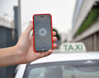 Someone holds a smartphone with an Uber app open on it. A taxi is seen in the background. 