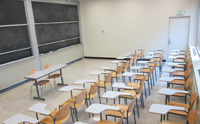 This small classroom, which can hold about 30 students, has three rows of chairs with attached desks. At the front, there is a small table with two chairs and two columns of sliding chalkboards.