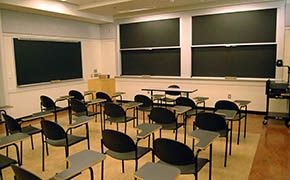 Four rows of tablet armchairs facing sliding chalkboards at front and side of room. An instructors table is in front of the chalkboards.