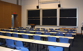 Classroom with 5 long tables and blue chairs on wheels; 3 sliding blackboards at the front of the room.