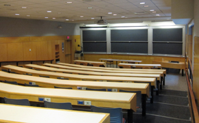 Classroom with even rows of long tables, each with about nine chairs. Six chalkboards at the front; projector mounted on the ceiling.
