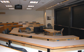 A large, modern classroom with faceted tiered seating, blackboards, and a projector.
