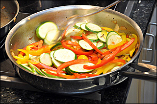 Zucchini and red and yellow peppers are being sautéed in a metal frying pan.