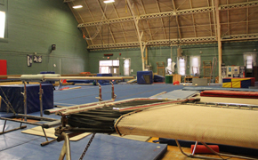 Gymnastics gymnasium featuring a trampoline and parallel bars in the foreground, a large blue practice mat in the middle of the space, and various incline mats and equipment in the background.