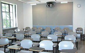 Twenty-nine light blue tablet chairs arranged in 6 rows. They are facing blackboards not visible in the picture. A large window is to the left of the chairs. A clock is on the back wall of the classroom. A projector is mounted to the ceiling.