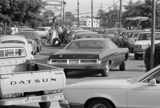 A black and white photo of cars and trucks lined up, surrounding a single gas pump.