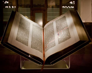 A large book, placed on a Plexiglas stand, lies open to a page containing elaborate type.