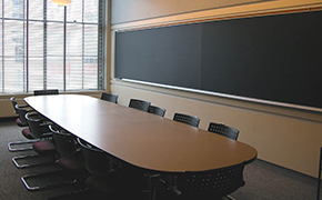 A small classroom with a large wood table surrounded by grey chairs, with chalkboards on either side.