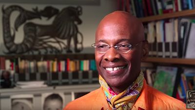 Smiling man wearing orange shirt, colorful scarf, and glasses. Bookshelves and file cabinets in the background.