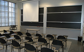 Classroom with about 20 tablet desks arranged in 4 rows. An instructors table is at the front of the room. A white projection screen is pulled down in front of a blackboard behind the instructors table.