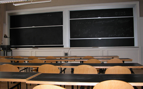 A classroom with a large sliding chalkboard at the front and rows of tables and chairs for students.