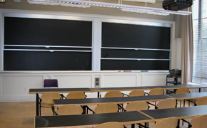 This classroom contains rows of long tables, with each row having roughly six chairs behind it. At the front, there are two columns of sliding chalkboards.