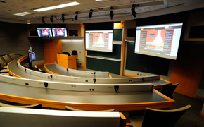 A classroom with chalkboards, podium, slide screen at the front and tiered rows of seating for students.