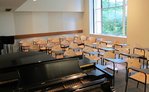 Photo of classroom showing a grand piano and about 25 movable chairs/desks in rows.