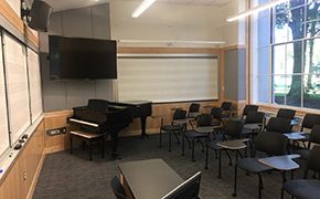 Photograph of classroom with a piano on the far wall, white boards on the far and left wall. Tablet chairs along the right wall of windows.