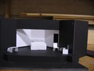 A small desk and chair sits stage right, while center stage is dominated by an office space with shelves, a large desk and chair, and a door leading offstage left. On the far left, two chairs and an endtable form a conversational grouping.