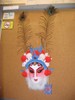 A dramatically painted white mask with red makeup accents on the cheeks, eyes, and lips wears a red, white, and blue crown surmounted by two peacock plumes, and large blue dangling earrings.