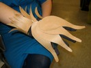Drumstick-shaped brown paper cutouts overlap on the back of the hand and curve down over each finger. The cuff is made of paper with random tapers and cuts, curving up from the arm.