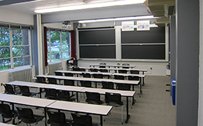 Photo of classroom with 5 wide tables in rows, seating 6 each, and a pair of chalkboards at the front of the room.