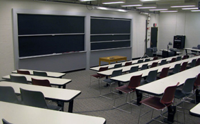Classroom with rectangular tables and chairs arranged in tiered rows. A small brown table is positioned at the front of the room. It sits in front of two chalkboards.