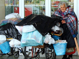 An elderly woman on crutches waits outside a train station with  bags, buckets, and blankets resting on two wheelchairs beside her.