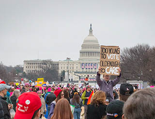 A photo of a young girl in a crowd of people holding up a sign that reads 'Too young to vote, old enough to care' during the 2017 Women's March on Washington.